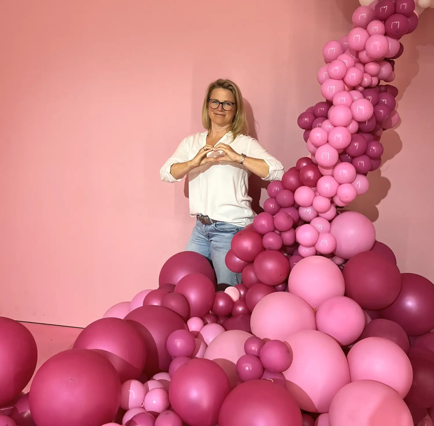 Kirsten Kau macht ein Handherz, während sie zwischen rosa Luftballons steht